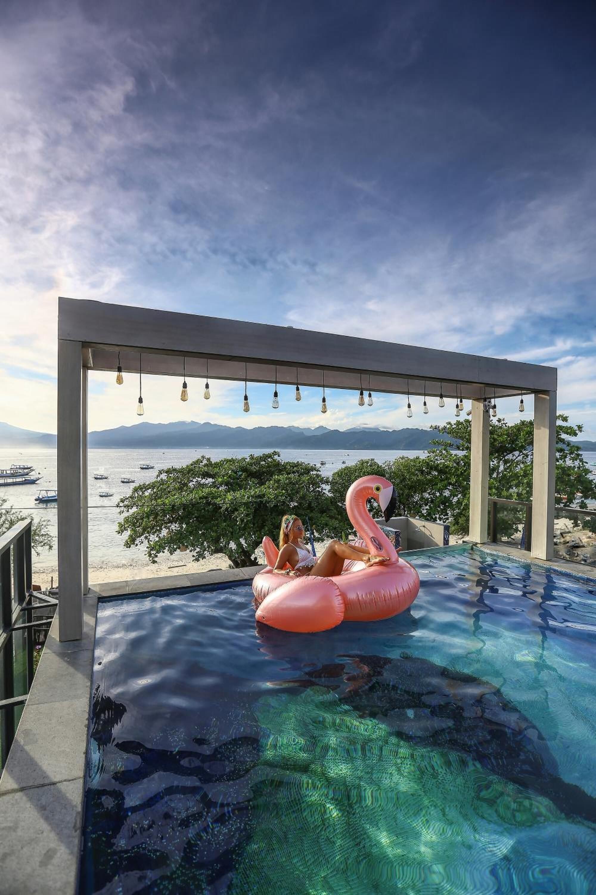A swimming pool with a pink flamingo float, surrounded by a pergola with hanging lights, overlooking a body of water and distant mountains.
