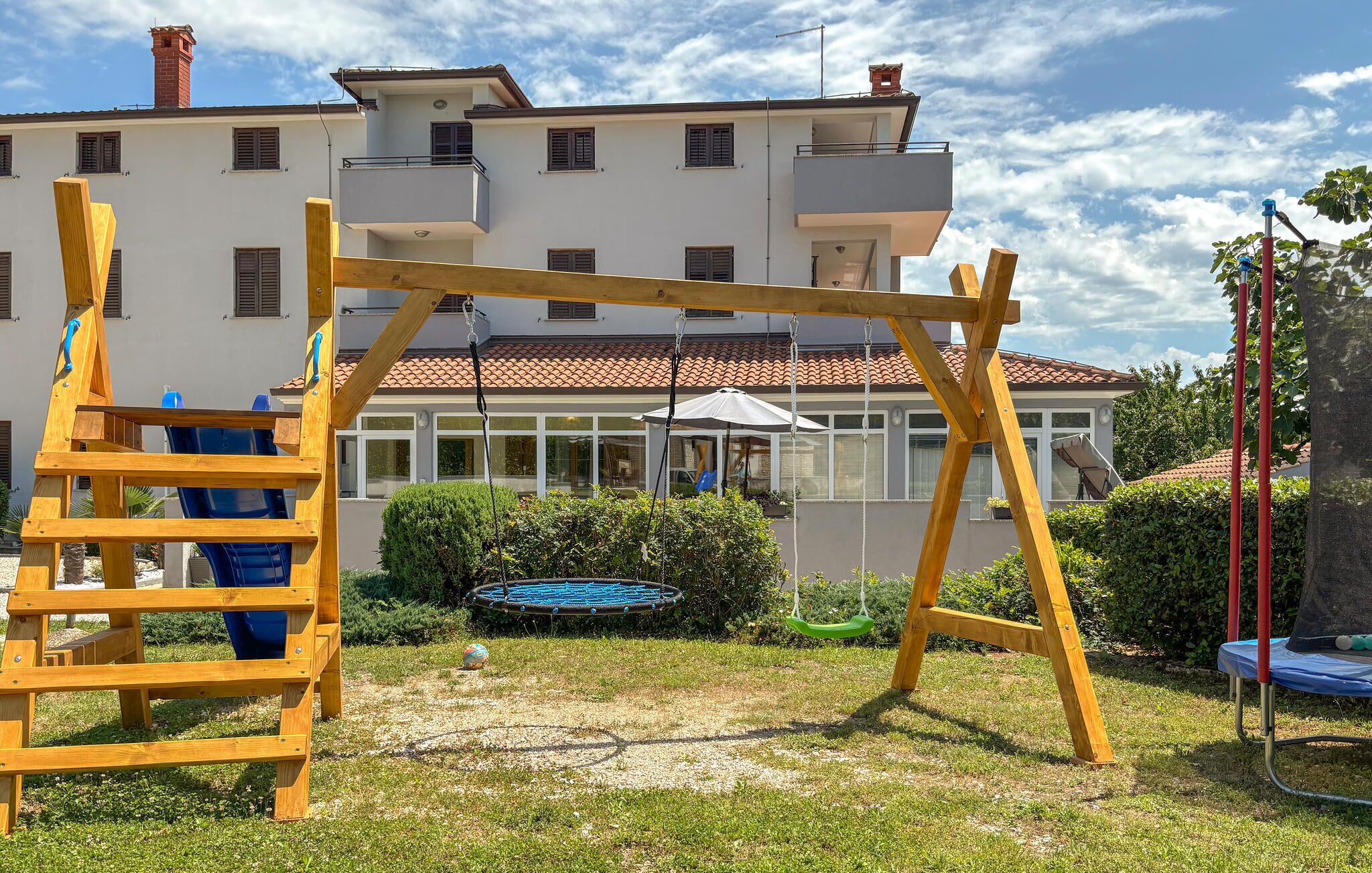 Un parc infantil amb un joc de cadira de balanç de fusta i un tobogan, un trampolí i una casa al fons.