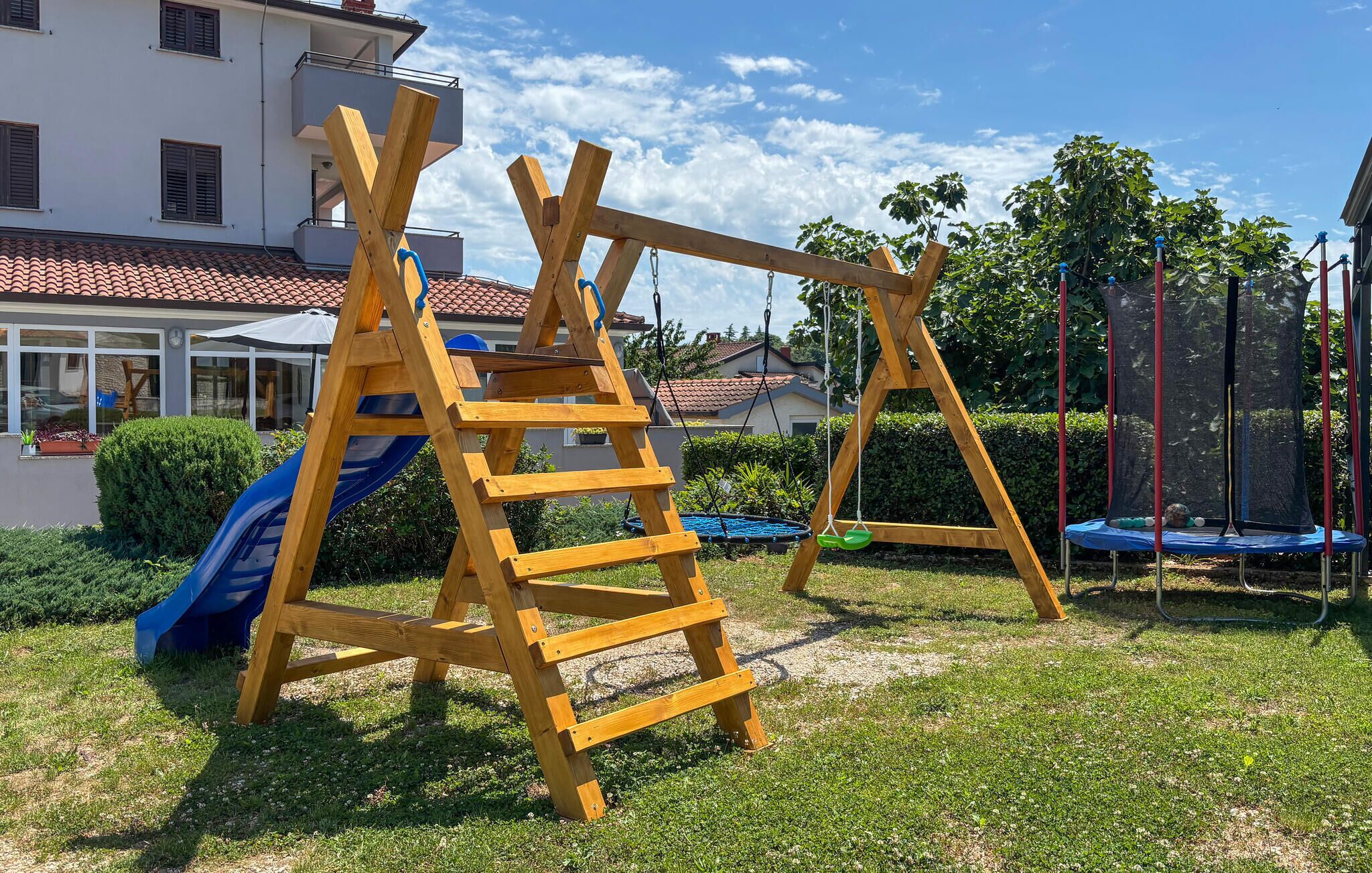 A wooden swing set and slide in a grassy yard with a house in the background.