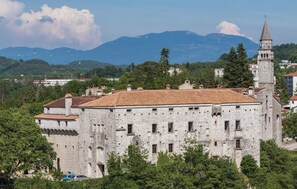 A historic stone building with a tiled roof, surrounded by greenery and a mountainous backdrop.