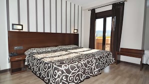 A hotel room with a large bed, a nightstand, a desk with a telephone, a bench, and a view of the outdoors through a window. - Hotel Ciudad de Plasencia (Plasencia)