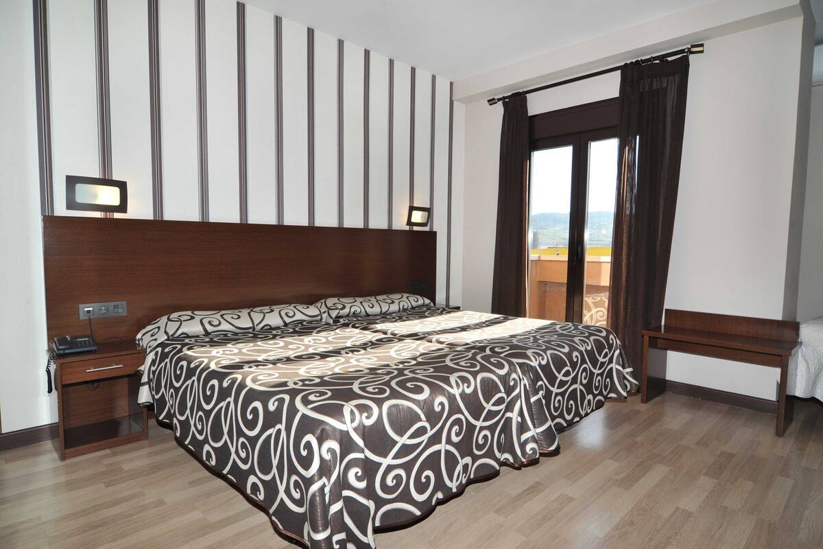 A hotel room with a large bed, a nightstand, a desk with a telephone, a bench, and a view of the outdoors through a window.