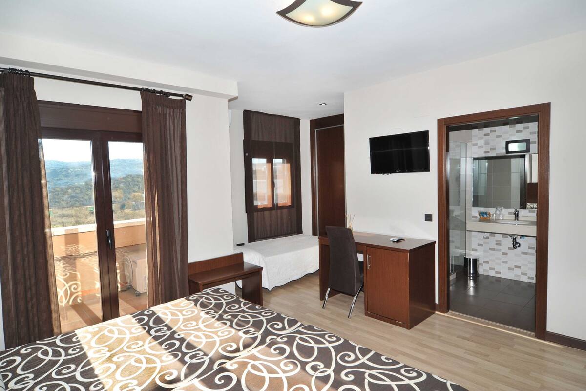 A hotel room with a bed, desk, chair, and a balcony with curtains.
