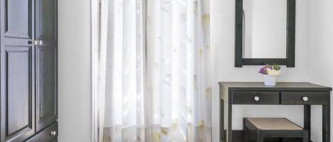 Una habitación con un armario de madera oscura, una cortina blanca con estampado de pájaros, una mesita con espejo y un banco.