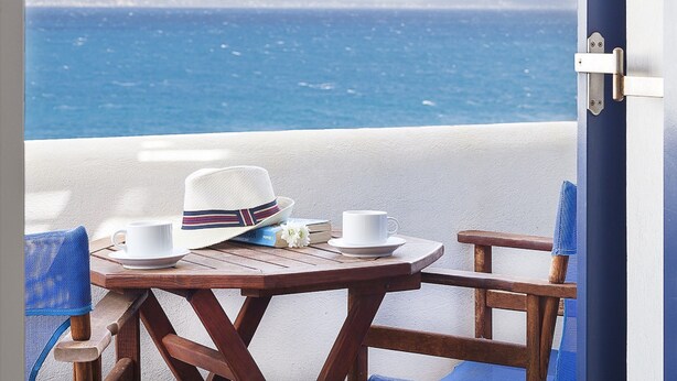 A wooden table with a white cup, a white saucer, a striped hat, and a book on a balcony overlooking the sea. - San Antonio Summer House by GHH (Paros)