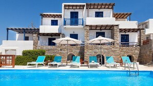A poolside area with lounge chairs and umbrellas, a stone building with multiple balconies, and a clear blue sky. - San Antonio Summer House by GHH (Paros)