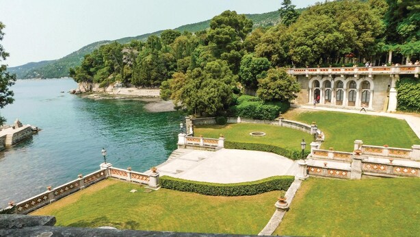 Un jardín ajardinado con un estanque, un edificio formal con ventanas de arco y un paisaje montañoso al fondo.