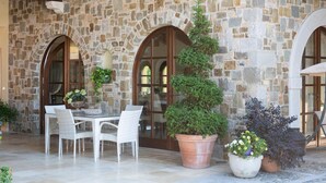 Un edificio de piedra con ventanales arqueados y un patio con mobiliario de mimbre blanco y plantas en macetas.
