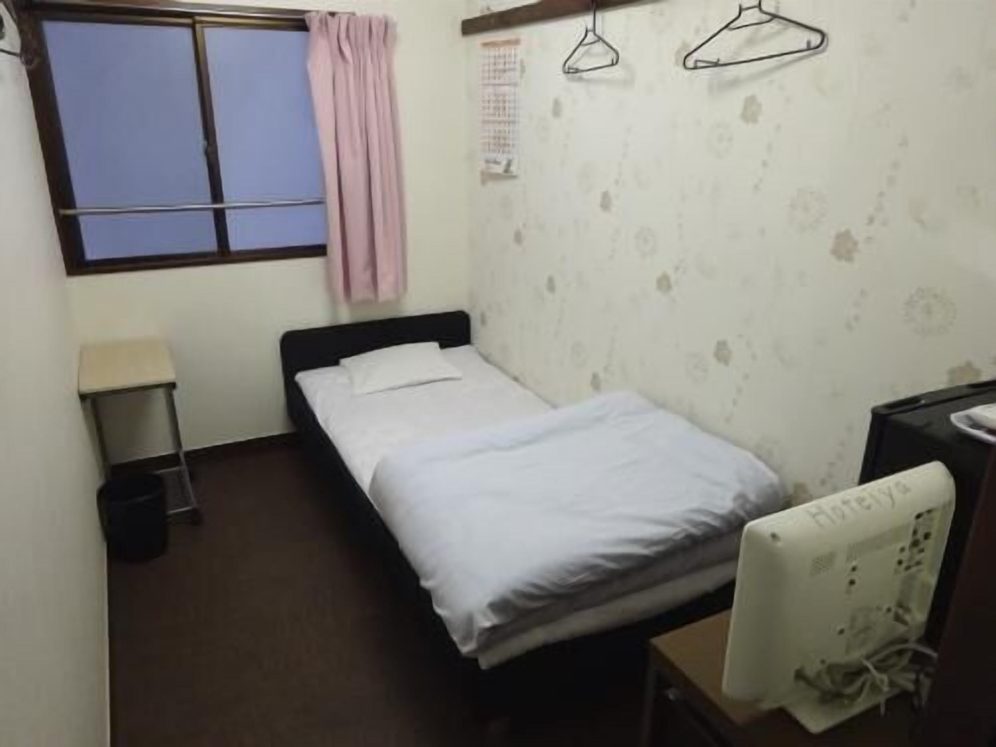 A single bed with white bedding, a bedside table, a small trash bin, a window with a curtain, and a wall-mounted heater.