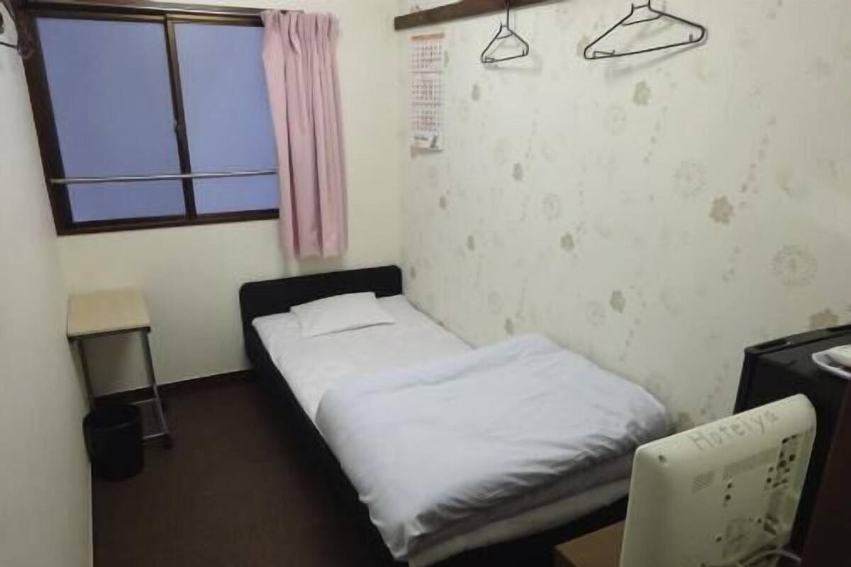 A single bed with white bedding, a bedside table, a small trash bin, a window with a curtain, and a wall-mounted heater.