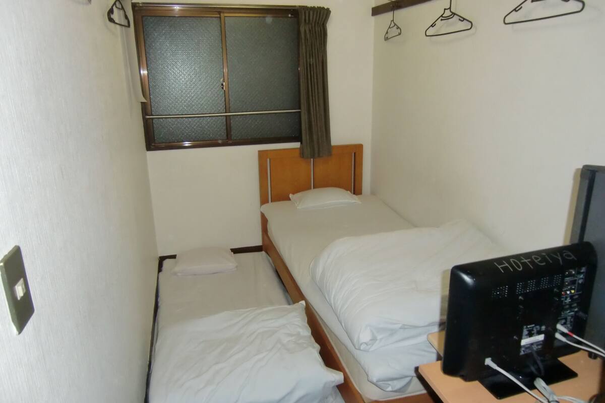 A small hotel room with two single beds, a window with curtains, a desk with a computer, and a wall-mounted TV.