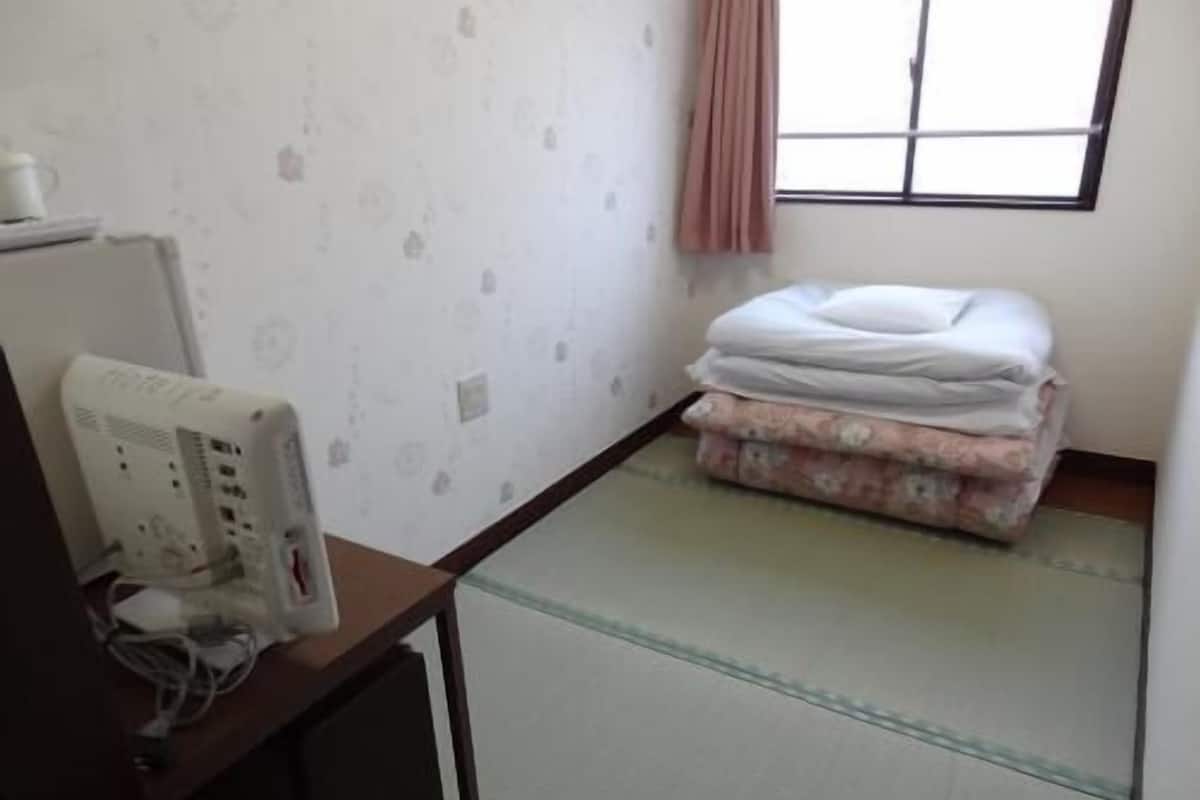 Una habitación con suelo de estera de tatami, cama con colcha floral, mesita de noche con un dispositivo blanco y ventana con cortina.