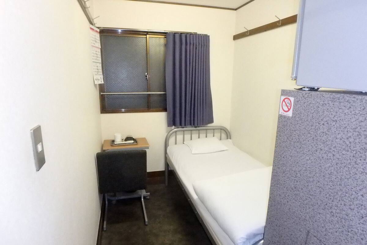 A small, compact room with a single bed, a desk with a chair, a small table with a cup and a phone, a window with curtains, and a no-smoking sign.