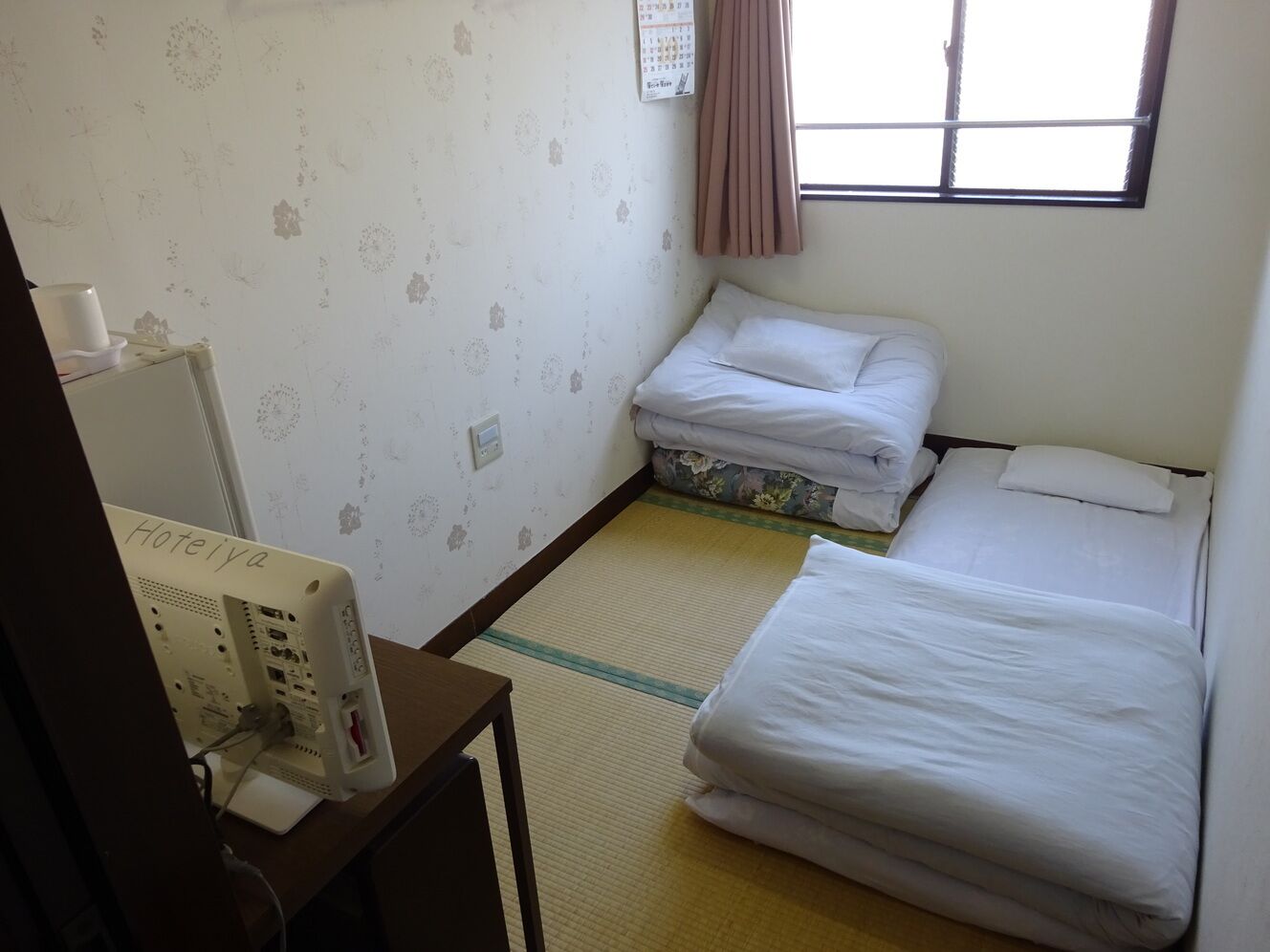 Une chambre d’hôtel avec deux lits, un bureau avec un ordinateur, un calendrier accroché au mur et une fenêtre avec des rideaux.