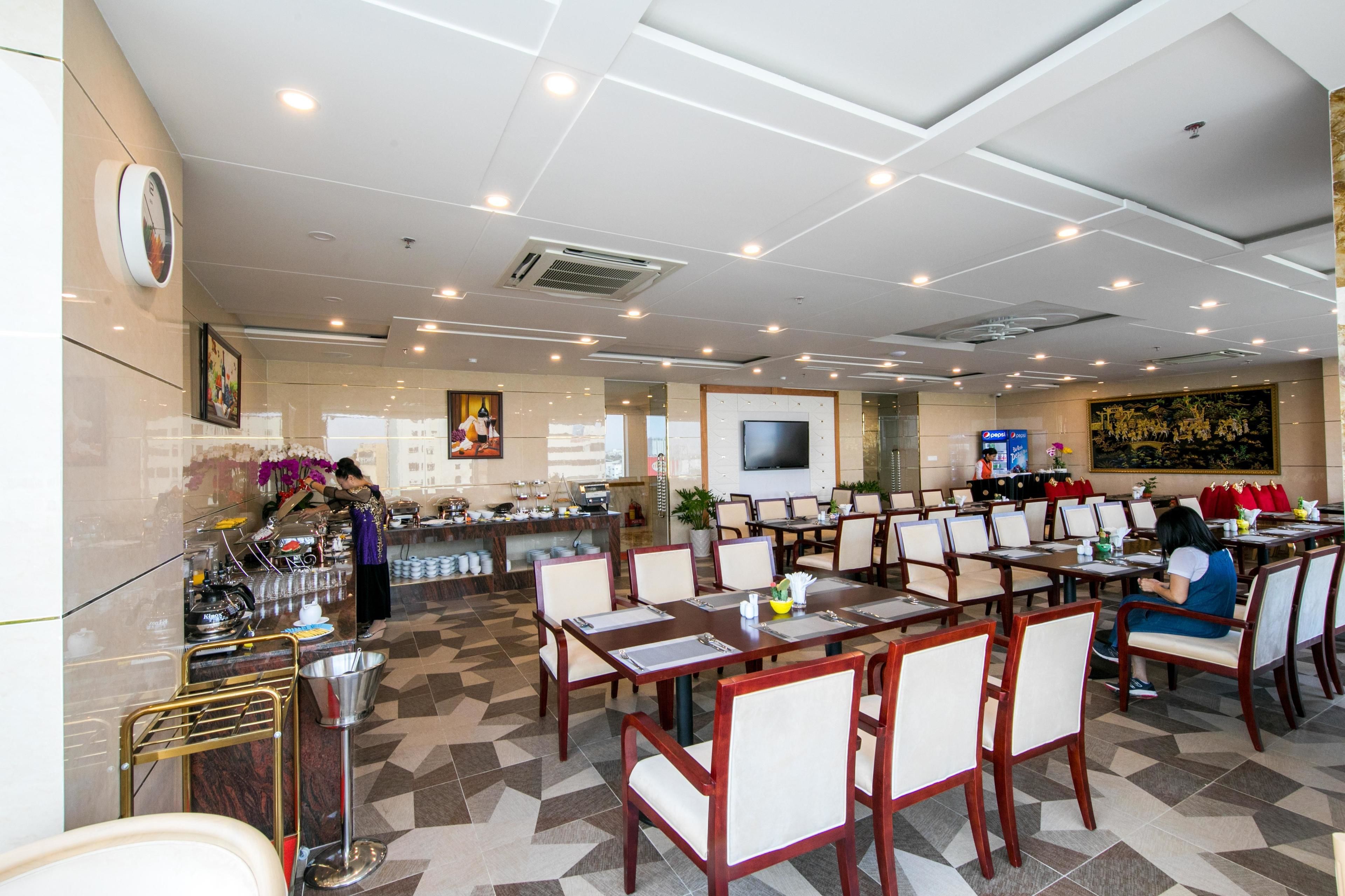 A spacious restaurant with multiple dining tables and chairs, a kitchen area, and a TV mounted on the wall.