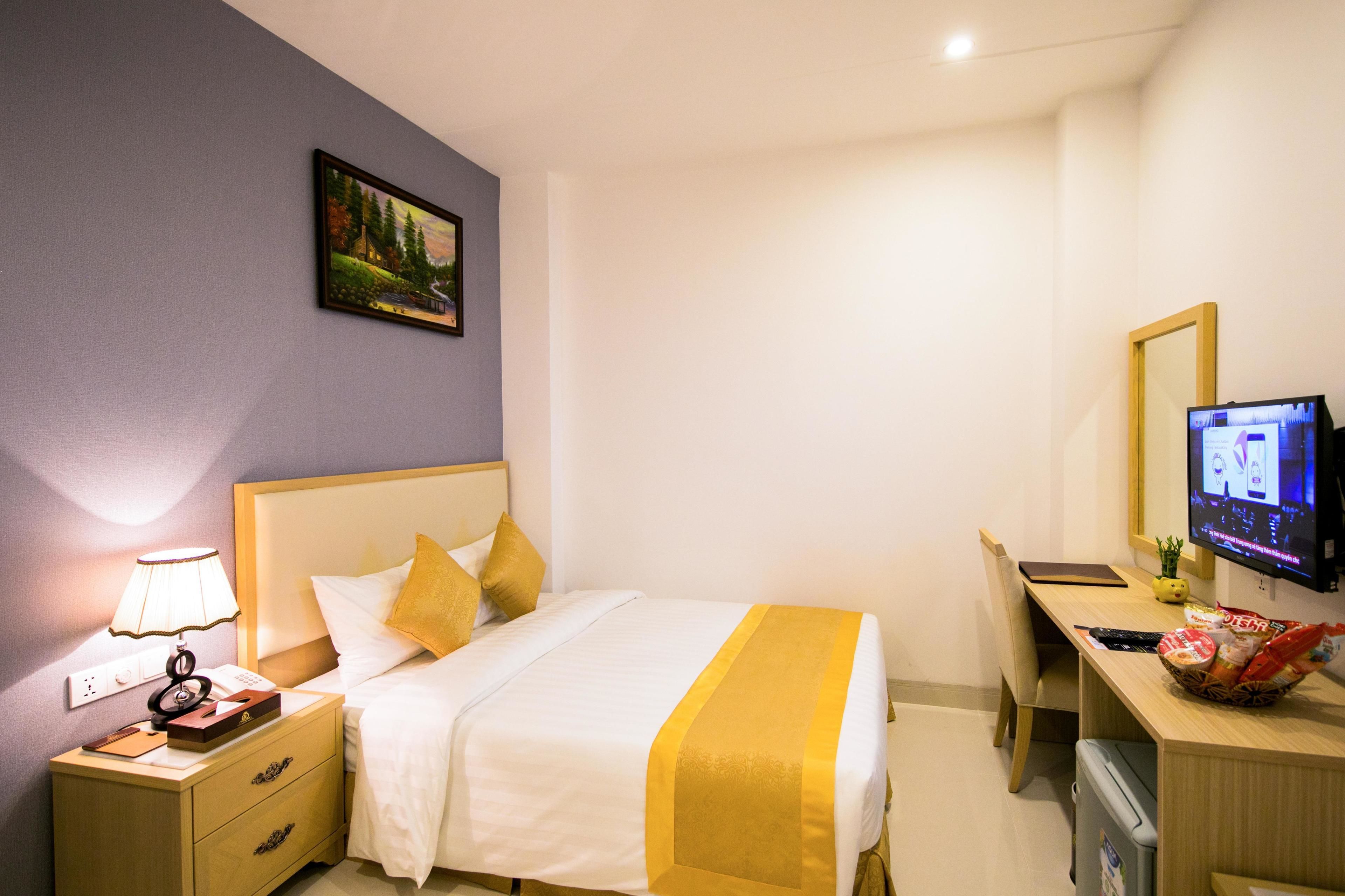 Quarto de hotel com cama, mesa de cabeceira, luminária, televisão, escrivaninha e um pequeno cesto de lixo.