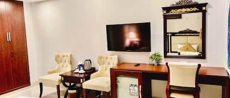 Habitación de hotel con televisor de pantalla plana grande, escritorio de madera, dos sillones, refrigerador pequeño y espejo con marco decorativo.
