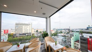 Hotelkamer met een groot raam dat uitzicht geeft op de stad, inclusief een hoog gebouw en een weg met voertuigen. Er zijn verschillende stoelen en tafels klaar gezet voor zitplaatsen.