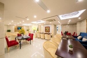 A modern hotel lobby with a central dining area, comfortable seating, and a large TV screen.