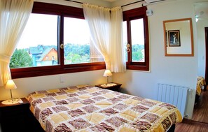 A bedroom with a bed, two bedside tables, a nightstand, a window with curtains, a radiator, and a framed picture on the wall.