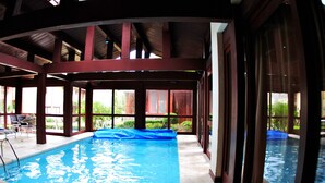 Una piscina interior con agua azul cristalina, rodeada por un espacio cubierto con vigas de madera y ventanas amplias.
