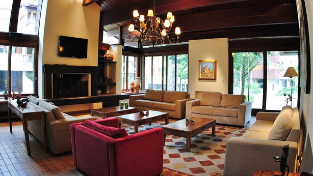Un salón amplio con una lámpara de araña grande, múltiples opciones de asiento que incluyen un sillón rojo, un sofá beige y un sillón a rayas. Un televisor de pantalla plana está montado en la pared, y hay una chimenea debajo.