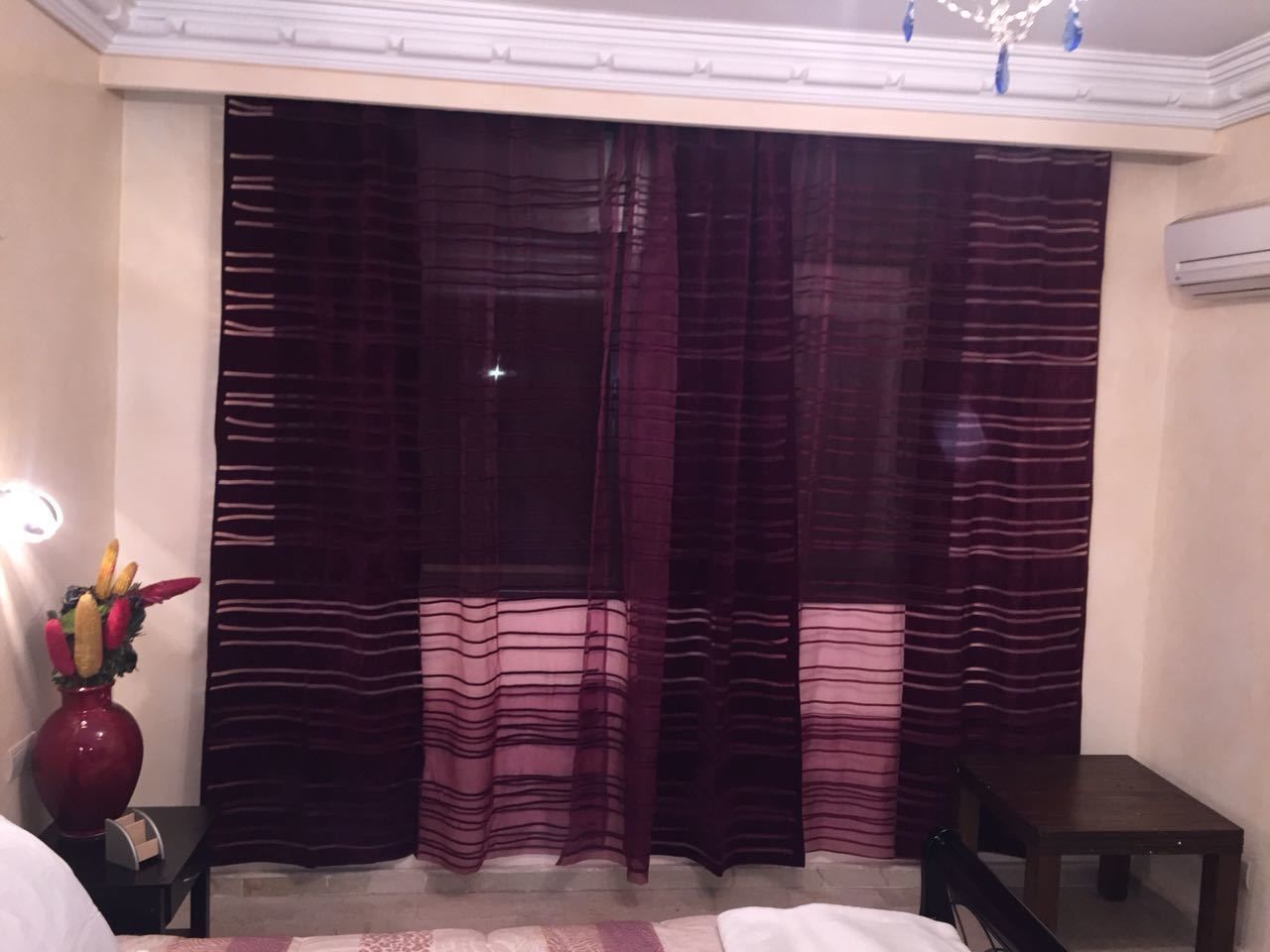 A hotel room with a bed, a nightstand with a vase of flowers, a small table, and a window with striped curtains.