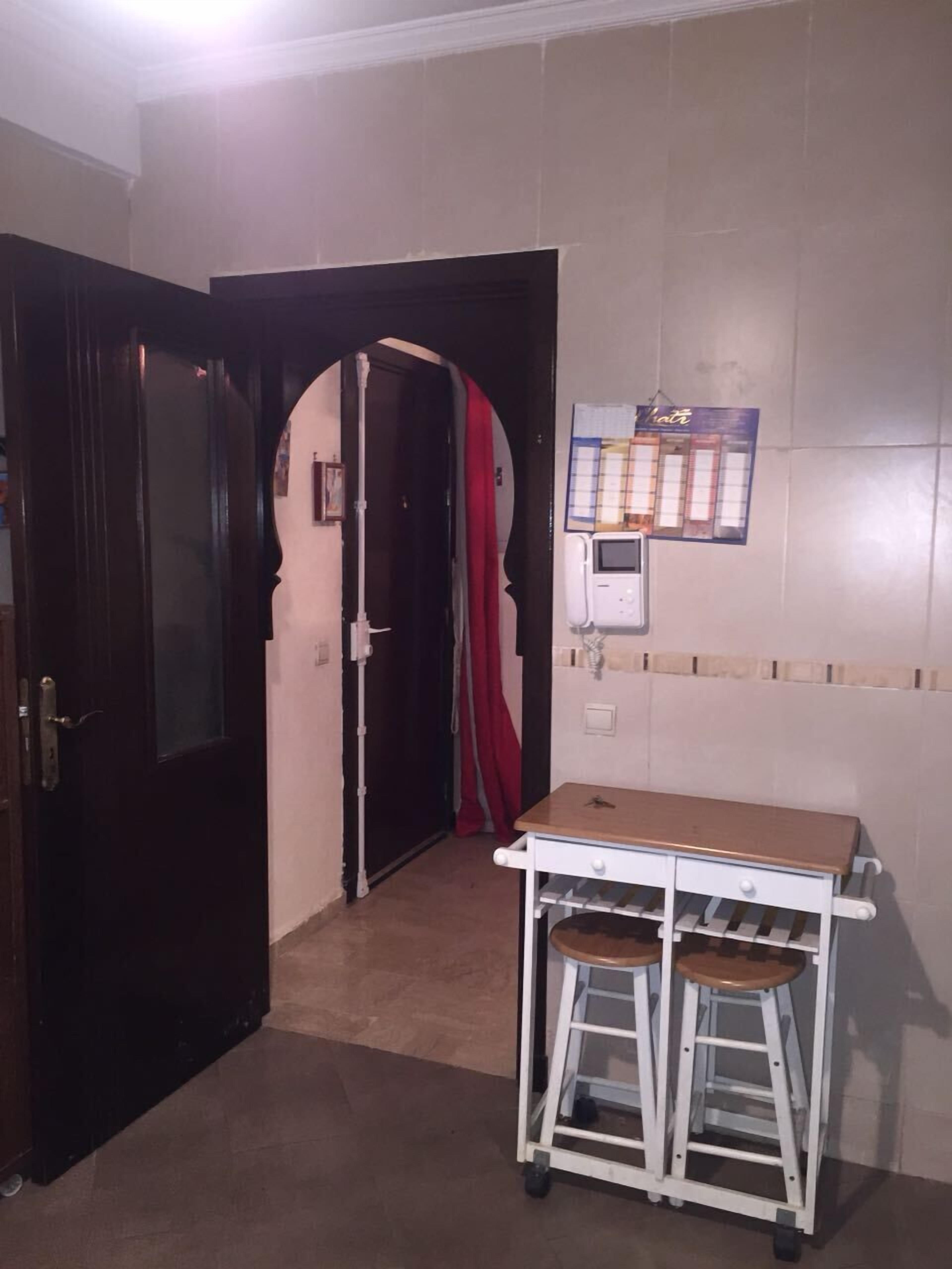 Una habitació amb una porta de fusta, una taula blanca i dos tamborets, i un quadre elèctric fixat a la paret.