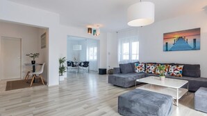 Un soggiorno moderno con un divano grigio, un tavolino da caffè bianco e un poggiapiedi grigio. Si nota anche un'area pranzo con tavolo e sedie, e una cucina sullo sfondo.