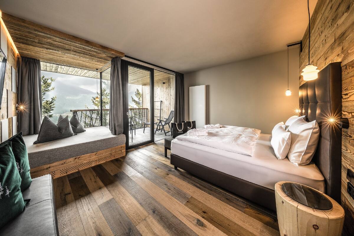 A bedroom with a bed, a bench, a view of the outdoors, and a wooden ceiling.