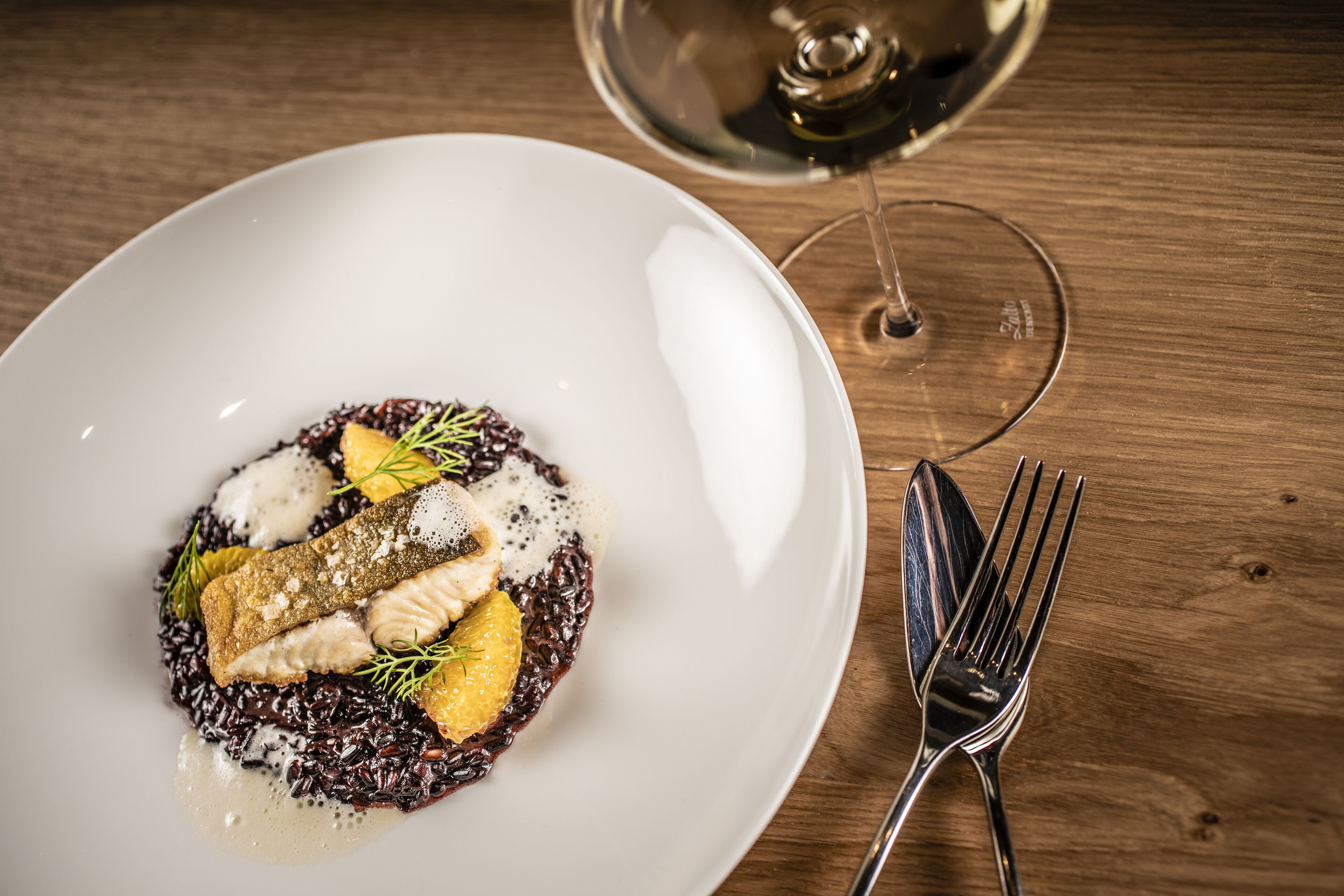 Un plato blanco con un guiso de pescado, adornado con hierbas y una rodaja de naranja, sobre una mesa de madera. También se ve una copa de vino y un tenedor.