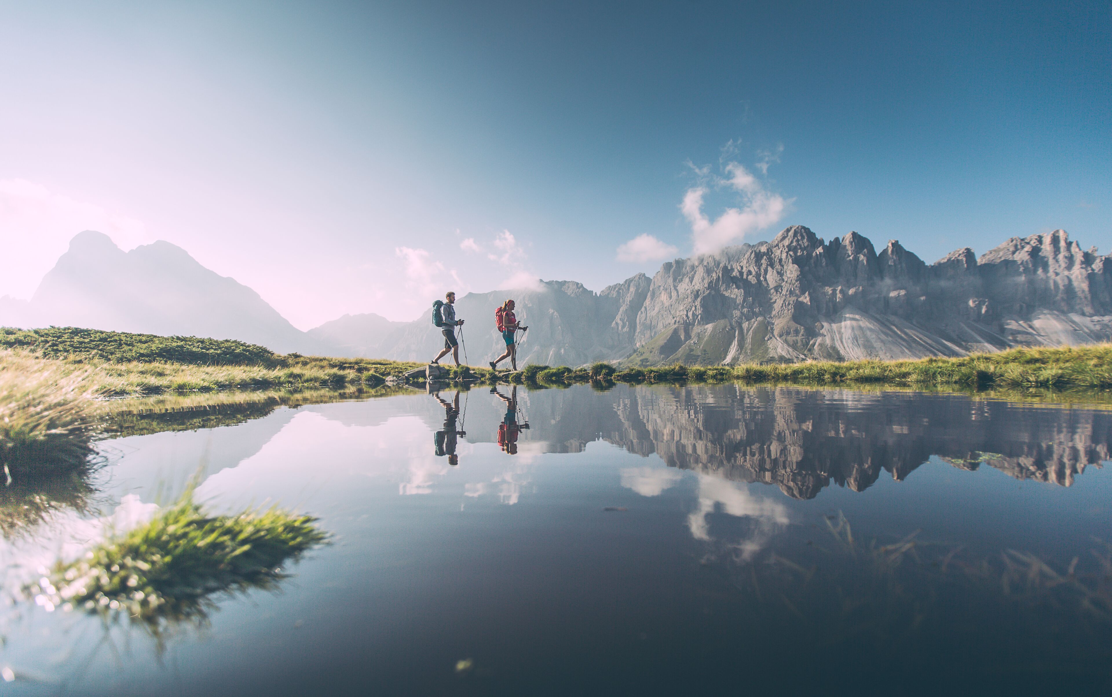 Ein ruhiger See mit deutlicher Spiegelung eines Wanderers und Bergen im Hintergrund.