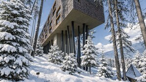 Un bâtiment moderne doté d’une structure en porte-à-faux originale, soutenue par des piliers, et entouré d’arbres couverts de neige, avec une chaîne de montagnes en arrière-plan.