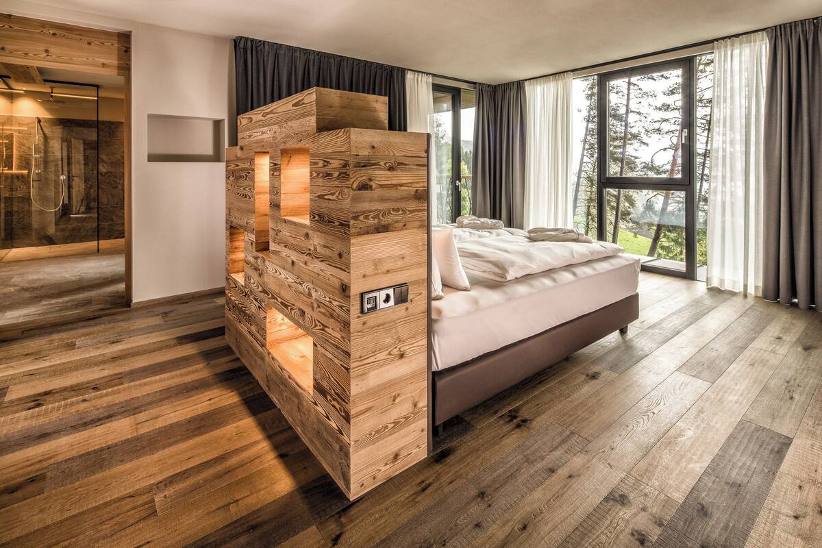 A modern bedroom with a wooden accent wall, a large window with a view of trees, and a separate shower area.