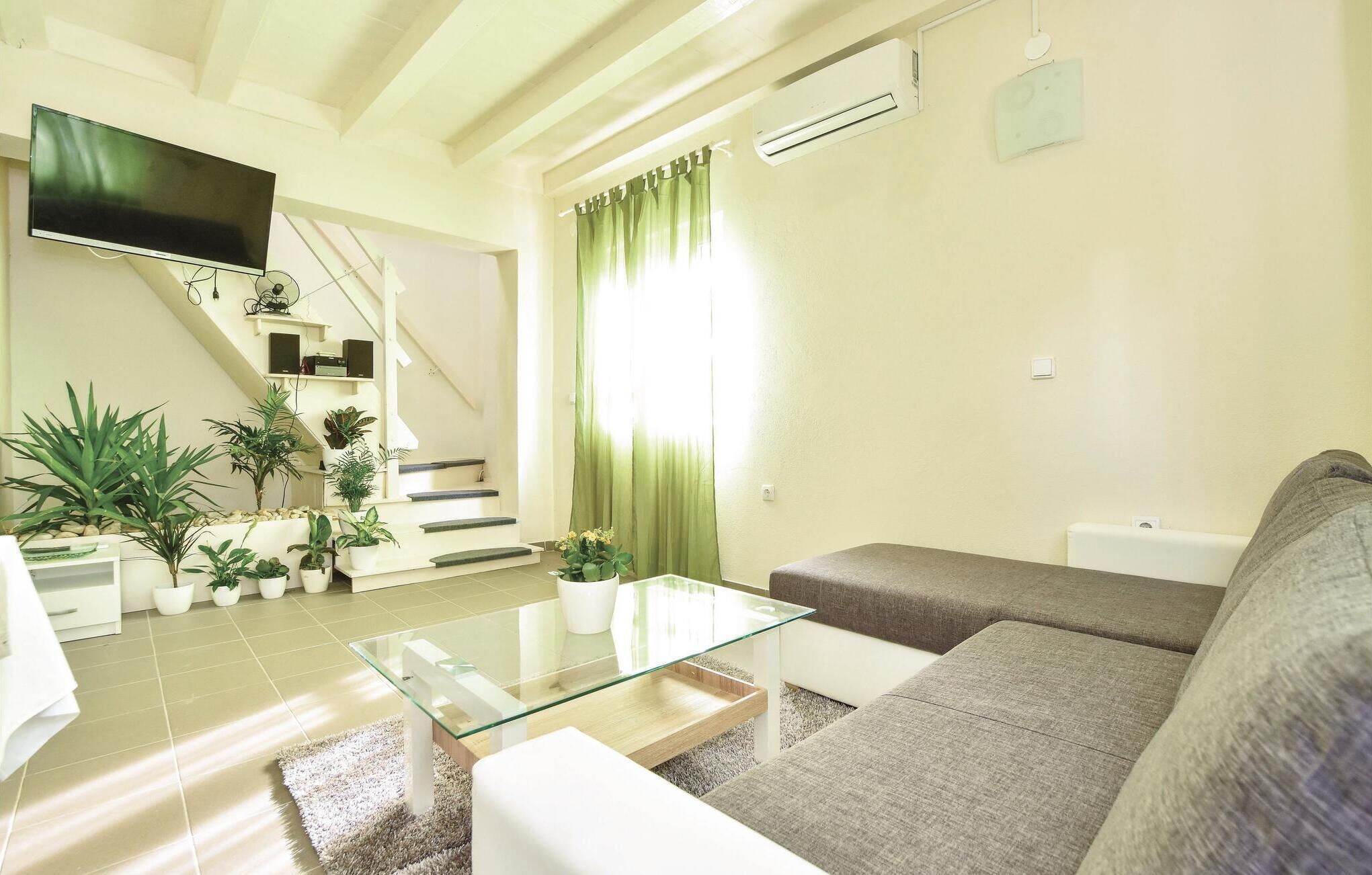 A modern living room with a glass coffee table, a sofa, and a TV mounted on the wall. There are potted plants and a staircase leading to another level.