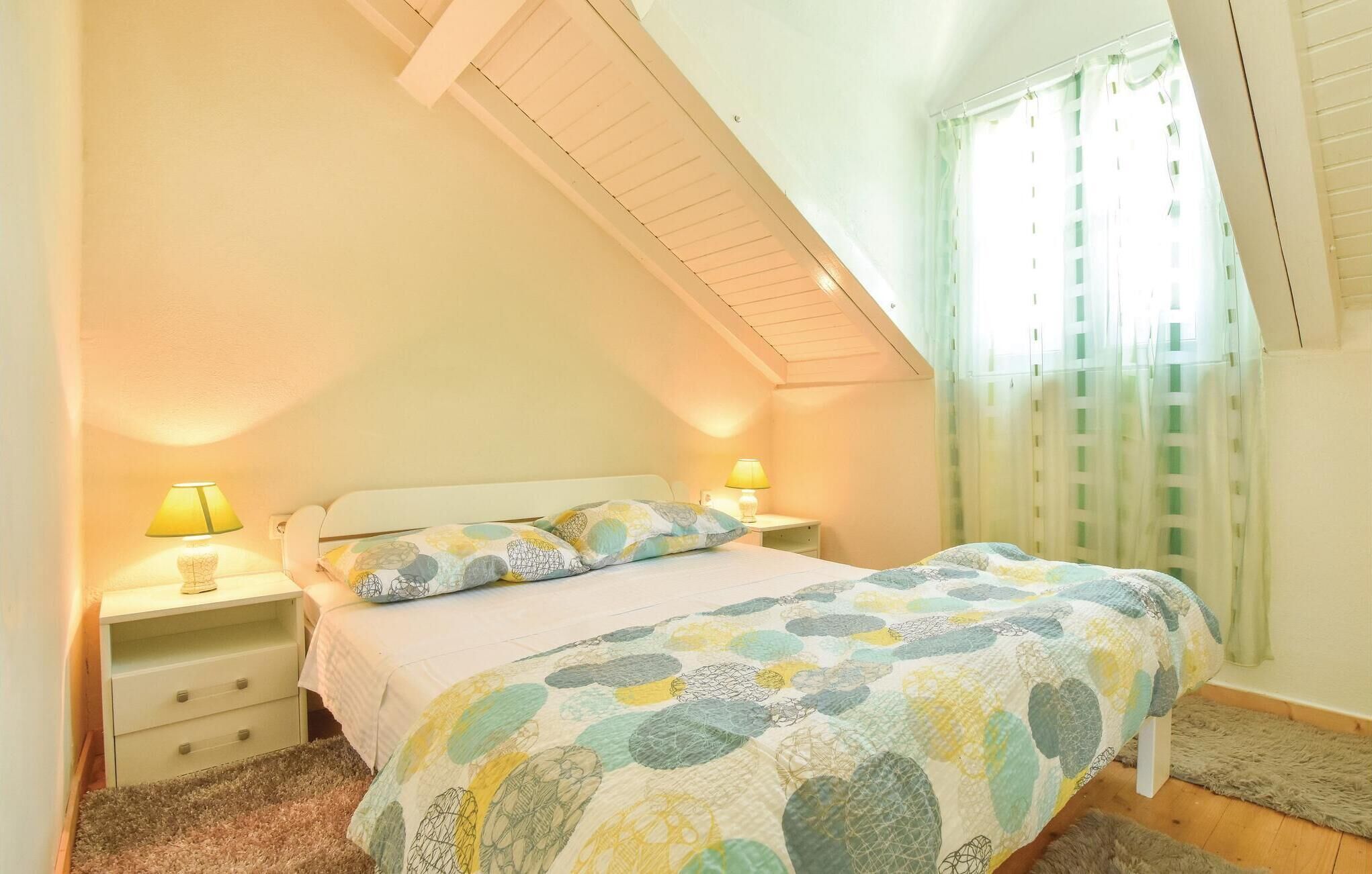 Un dormitorio con techo inclinado, cama con colcha estampada, dos lámparas de noche y una ventana con cortinas.