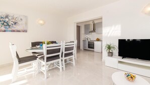 Een moderne, goed verlichte woonkamer met een eethoek, een keuken en een televisie op een witte kast.
