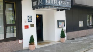 L’entrée d’un hôtel, avec une enseigne indiquant « HOTEL HOTELVERA ». Deux plantes en pot se trouvent de part et d’autre de l’entrée, ainsi qu’une porte et une fenêtre sur le côté gauche.