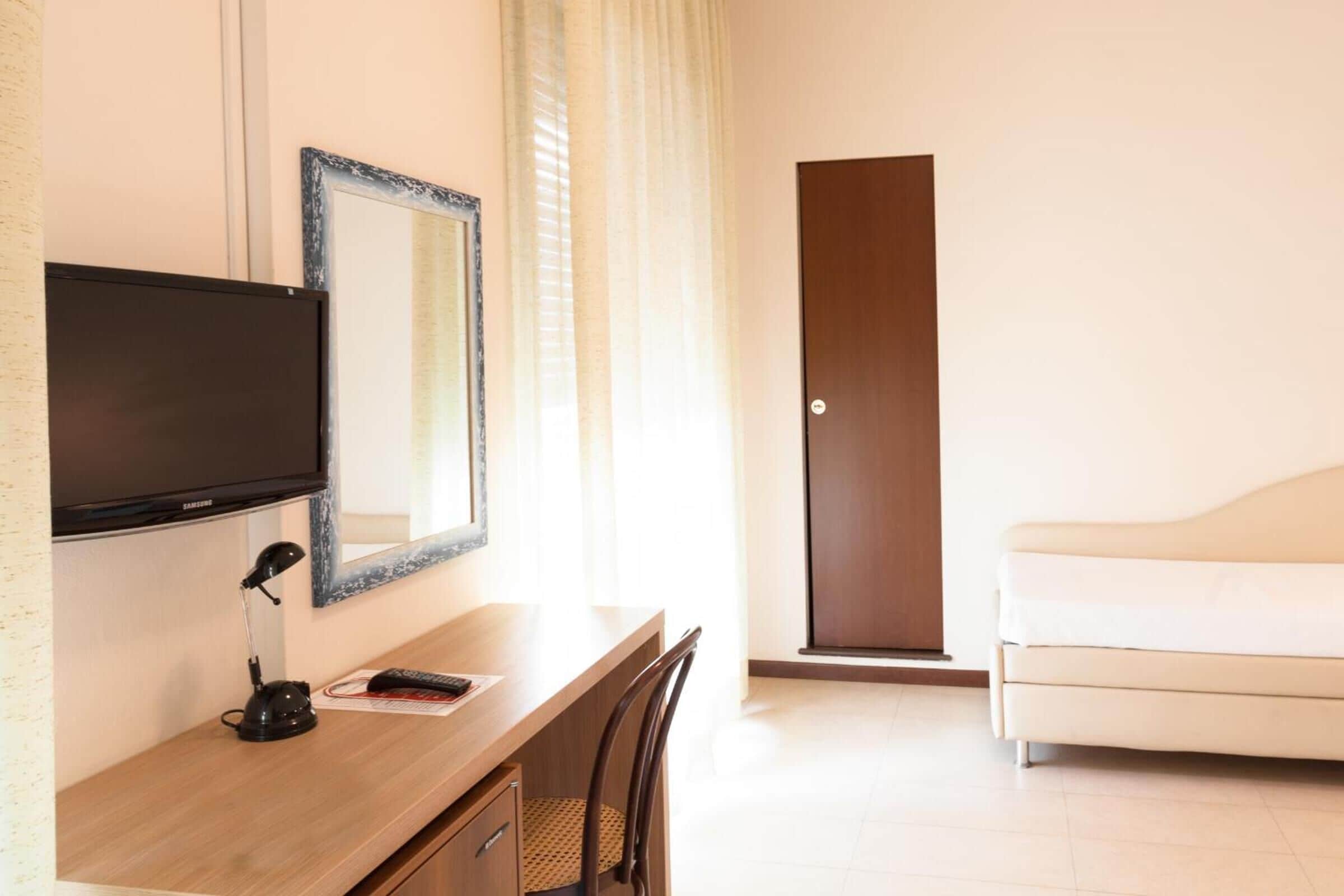 A hotel room with a wooden desk, a television, a mirror, a white sofa, and a door.