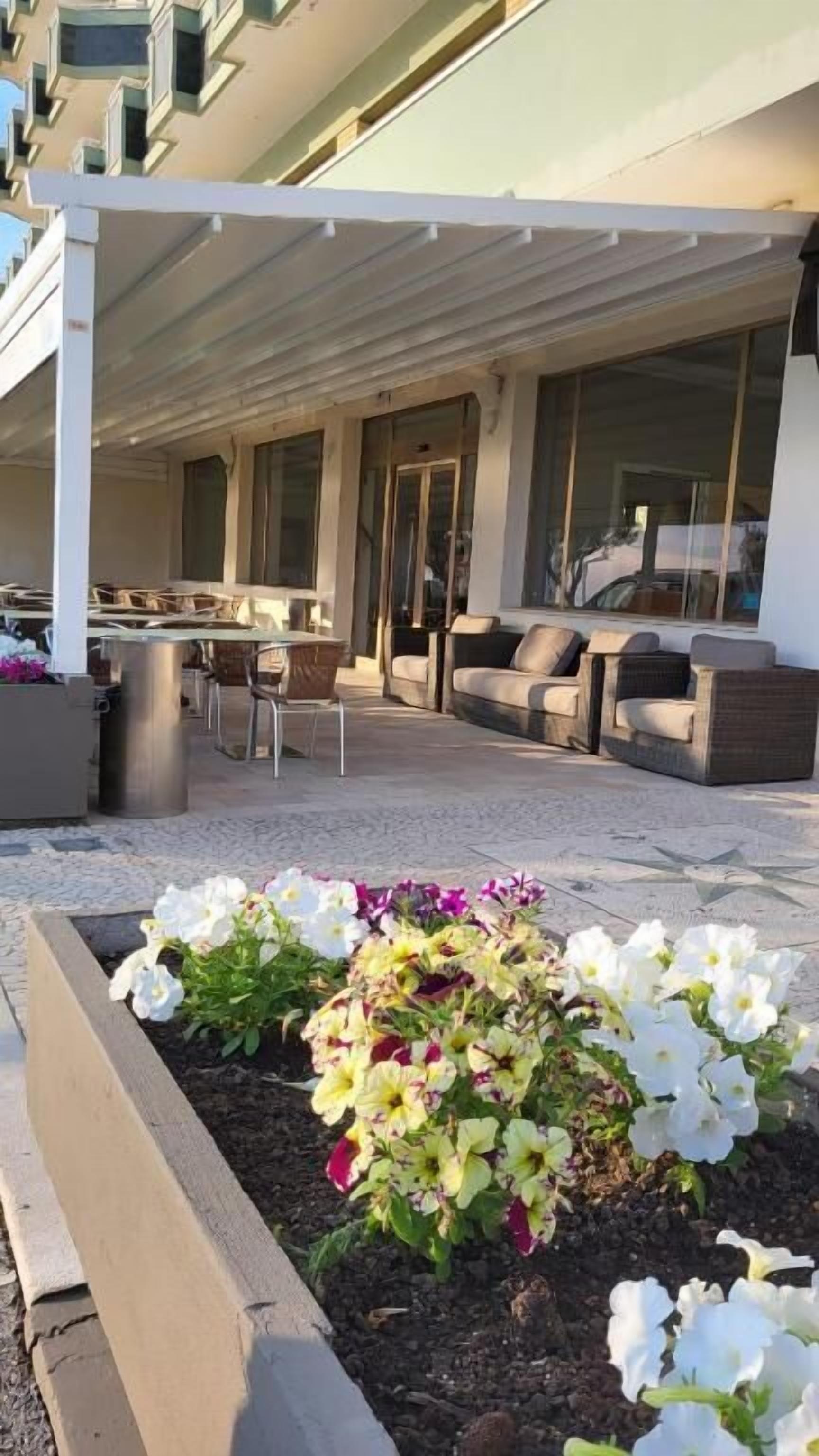 An outdoor seating area with a covered patio, cushioned seating, and a flower bed with white and yellow flowers.