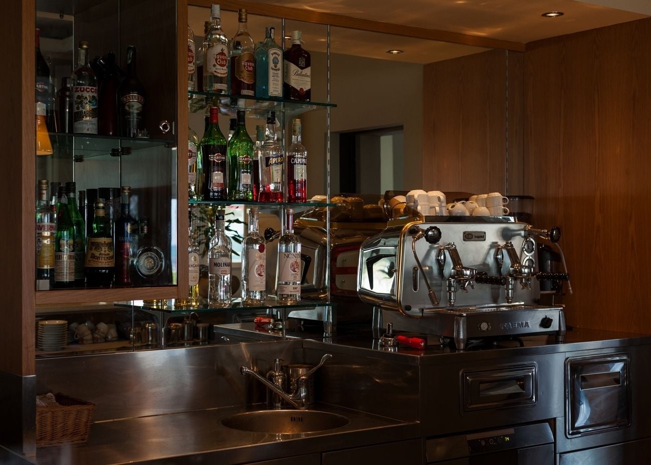 A bar area with a coffee machine, shelves of liquor bottles, and a sink.
