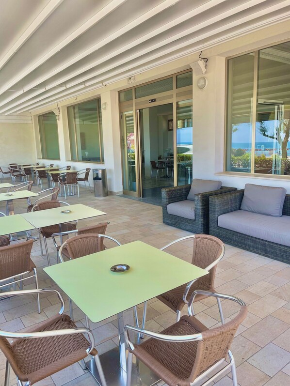 An outdoor cafe area with tables and wicker chairs, a sofa, and a view of the sea.