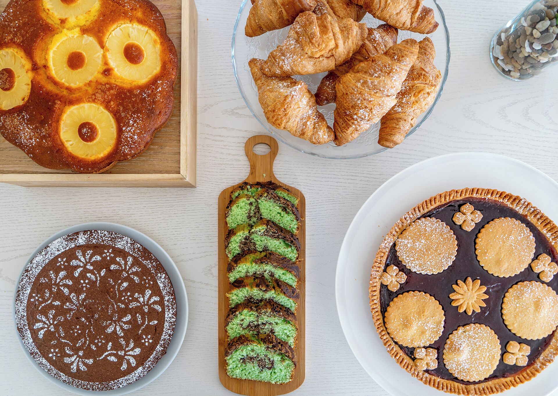 Variedad de postres, incluyendo pastel de piña, croissants, pastel de chocolate, pastel verde mármol y una tarta con galletas decorativas.