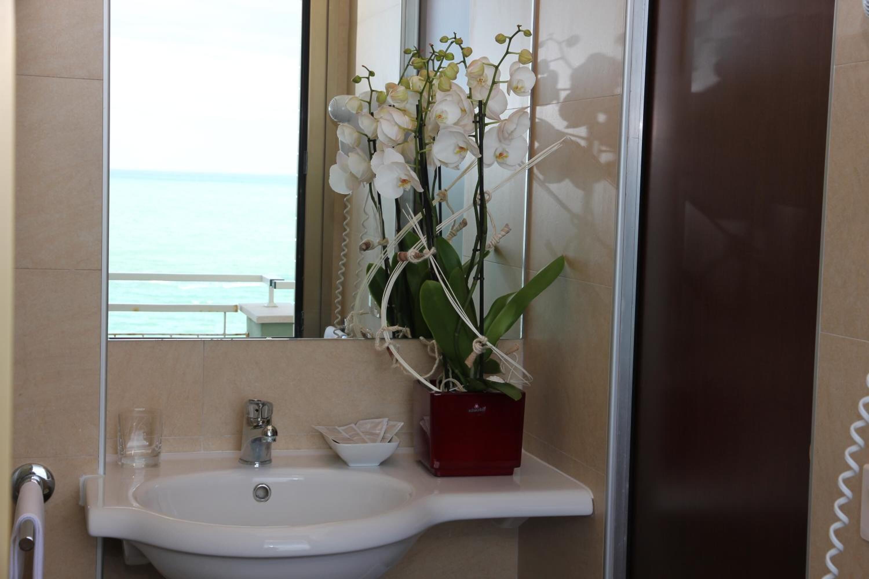 Un baño con un lavamanos blanco, un florero rojo con orquídeas blancas y un espejo grande que refleja el baño y una vista del mar.