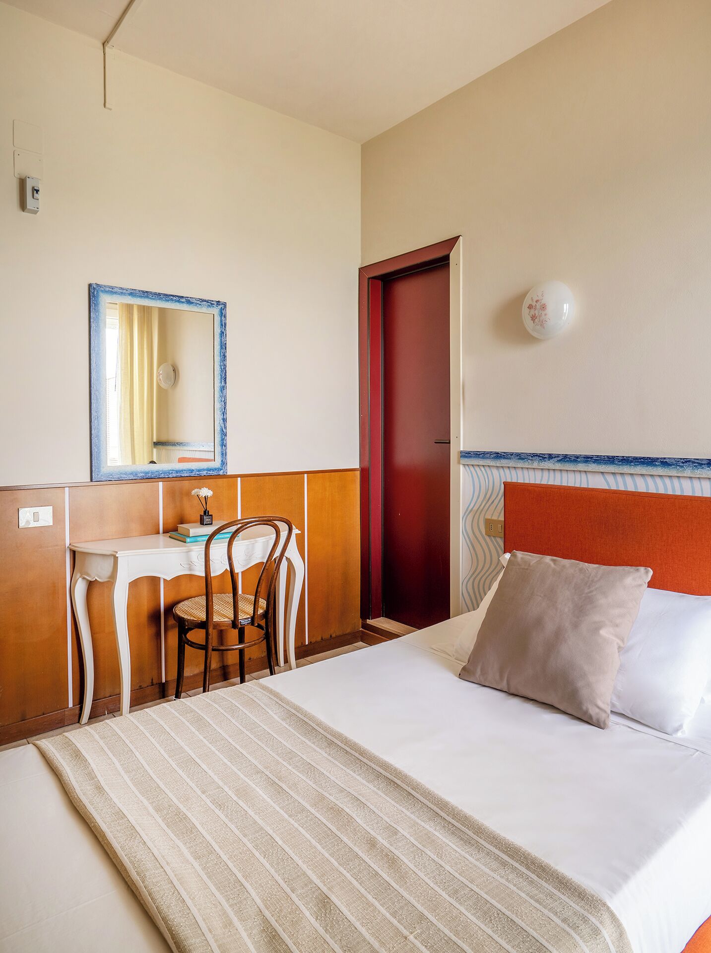 A hotel room with a bed, a desk, a chair, a mirror, and a door.