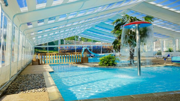 An indoor swimming pool with a water slide, a palm tree, and a covered area with a view of the outside. - Camping la kilienne (Warlincourt-lès-Pas)