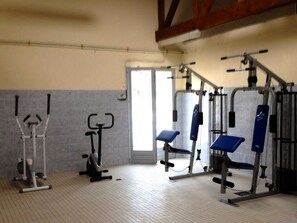 Een sportschool met trainingsapparatuur, waaronder een hometrainer, een multifunctioneel fitnessapparaat en een bank.
