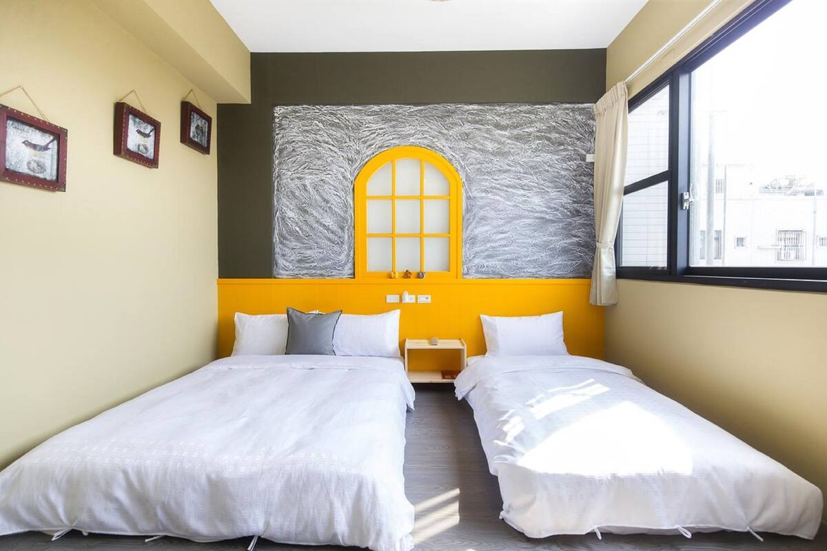 Habitación de hotel con dos camas individuales, un ventanal grande y un mural de un ventanal amarillo en la pared.