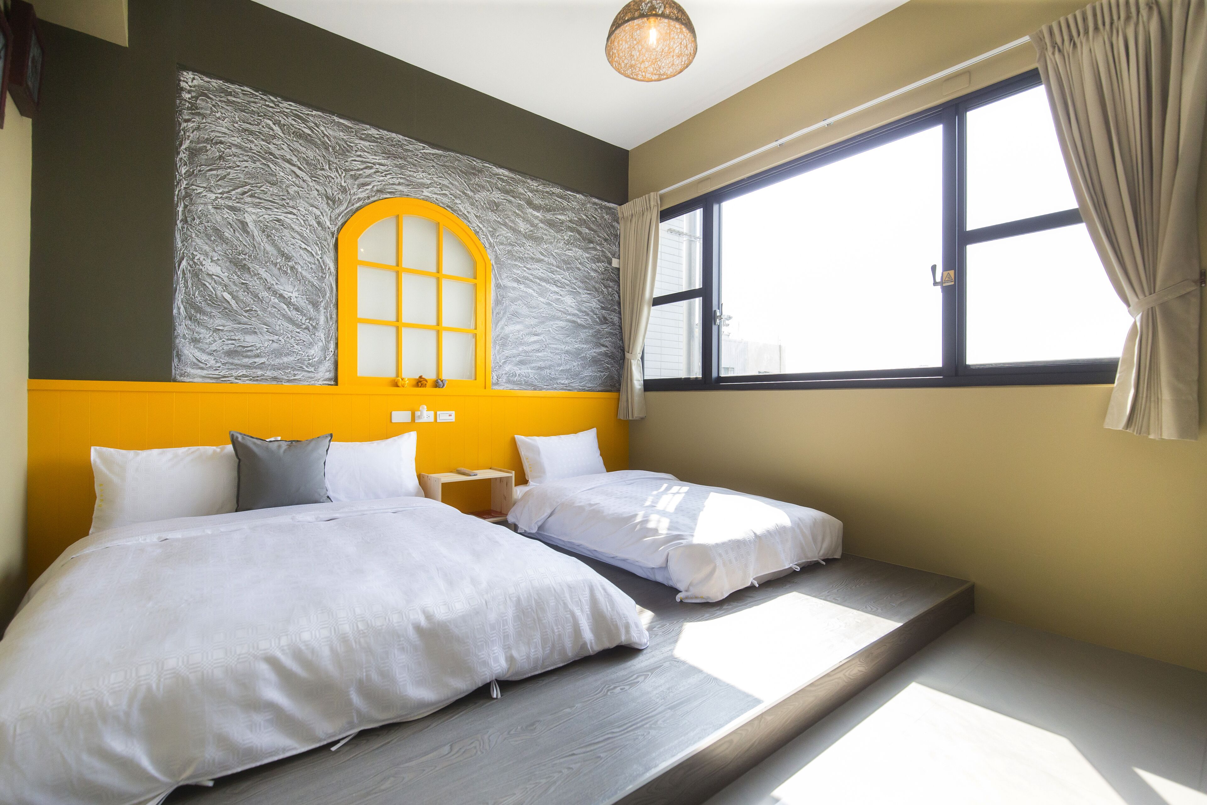 A bedroom with two beds, a window with curtains, a wall with a textured design, and a ceiling light.