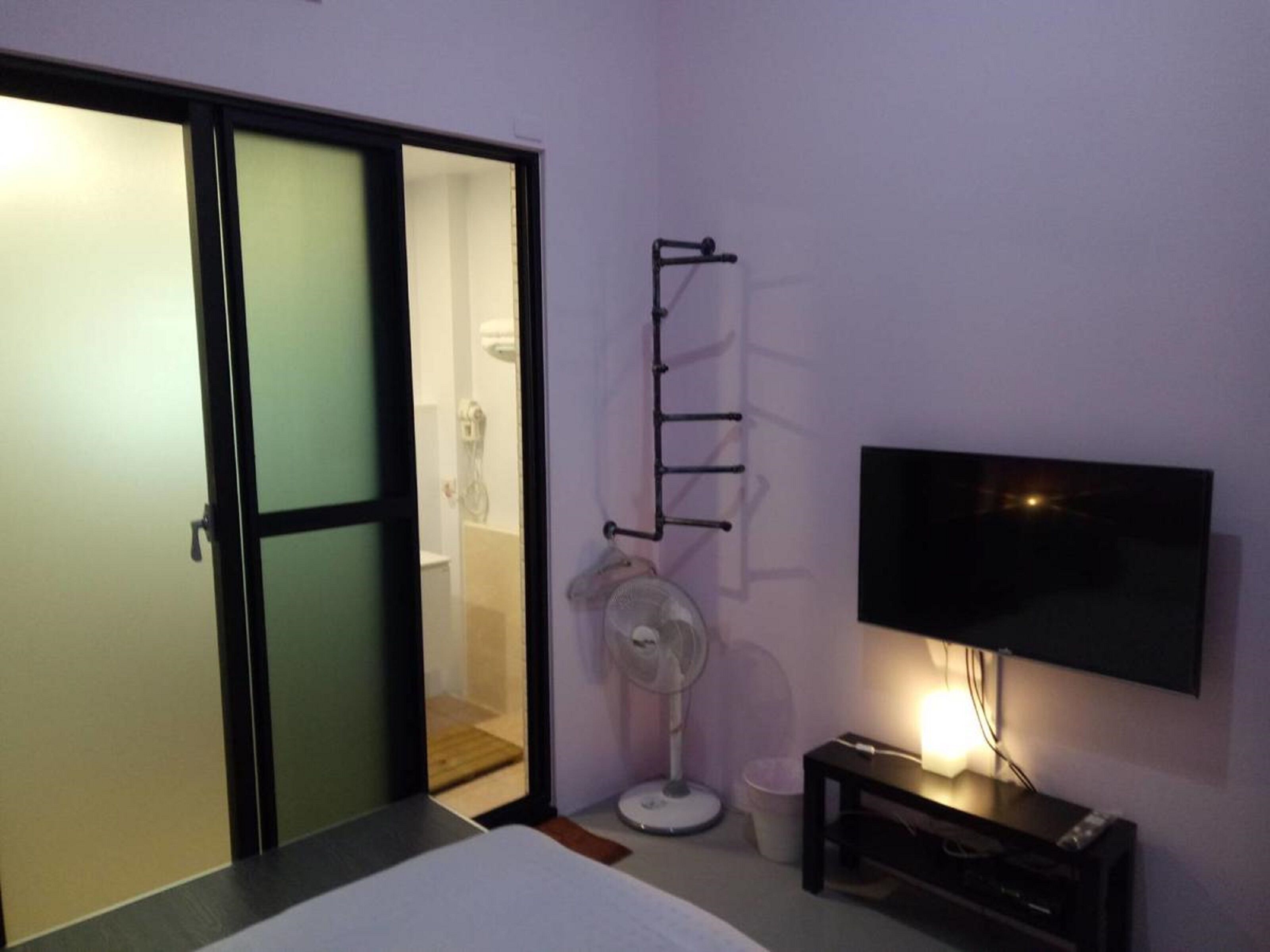 Una stanza con una porta scorrevole che dà su un bagno, una ventola a piantana, un televisore montato a parete e un comodino con una lampada.